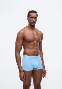 Μυώδης άνδρας όρθιος, φοράει ανοιχτόγαλανα εσώρουχα Calvin Klein boxer briefs, σε λευκό απλό φόντο με ουδέτερη έκφραση.