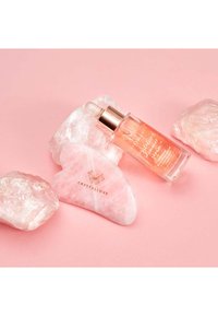 Strumento gua sha in quarzo rosa accanto a una bottiglia di vetro con contagocce contenente un siero rosa. Entrambi su uno sfondo rosa chiaro con due pezzi di cristallo grezzo.