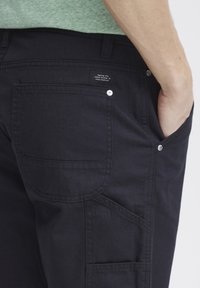 Pantalones cargo de algodón negros con un bolsillo trasero, costuras visibles y una etiqueta en la cinturilla; cuenta con cierres a presión y bolsillos laterales.