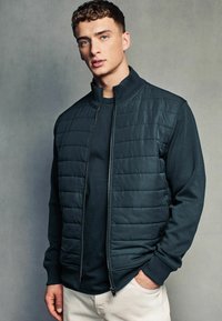 Gilet matelassé vert foncé avec col haut, fermeture éclair et manches longues sur un haut assorti de couleur foncée. Tissu lisse avec une texture subtile.