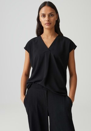 Femme aux longs cheveux foncés portant un haut noir ample à manches courtes et col en V ainsi qu'un pantalon noir texturé, les mains dans les poches.