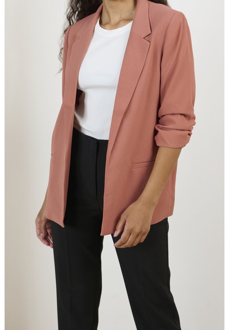 Prahově růžový blazer s klopami, ohrnutými rukávy a předními kapsami, kombinovaný s bílým ribbed topem a černými kalhotami.