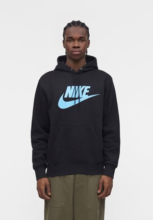 Giovane uomo con capelli intrecciati che indossa una felpa nera con cappuccio con un grande logo Nike azzurro chiaro, abbinata a pantaloni verde oliva, in piedi su sfondo bianco.