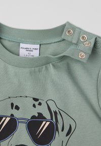 Vihreä puuvillainen t-paita, jossa on musta grafiikka aurinkolaseja pitävän koiran kuvalla. Olkapäällä on nepparikiinnitys, joka helpottaa pukemista.