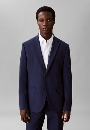 Młody mężczyzna w ciemnoniebieskim garniturze i białej koszuli stojący na tle jednolitego jasnoszarego tła, zwrócony przodem.