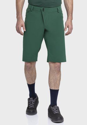Mann trägt grüne knielange Shorts, dunkle Socken und schwarze Turnschuhe, steht vor einem schlichten hellgrauen Hintergrund.