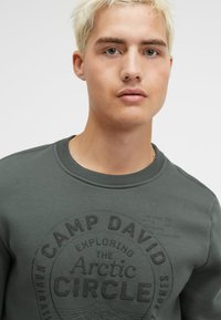 Dark grey sweatshirt with round neck, featuring black graphic text "CAMP DAVID" and "Arctic Circle." Made from soft, slightly textured fabric.