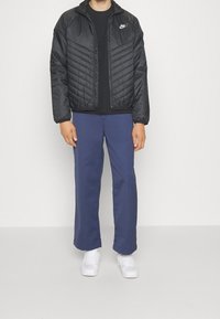 Černá prošívaná bunda se zipem, námořnické kalhoty a bílé tenisky. Bunda má vysoký límec a logo Nike na levém prsu.