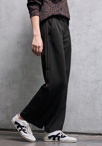 Pantaloni neri a gamba larga con vita elastica, caratterizzati da strisce laterali a contrasto. Abbinati a sneakers bianche con dettagli neri.