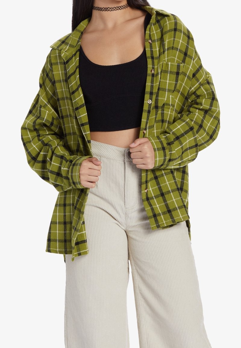 La personne porte une chemise à carreaux verte boutonnée ouverte sur un crop top noir et un pantalon en velours côtelé beige clair taille haute, tenant les bords de la chemise.