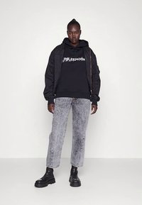 Casaco bomber preto com fecho éclair, capuz com texto gráfico e jeans cinzentos desbotados. Botas pretas robustas completam o conjunto.