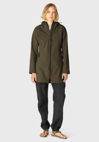 Chaqueta impermeable de un verde oliva oscuro con cremallera, dos bolsillos laterales y capucha, combinada con pantalones negros holgados y sandalias marrones.