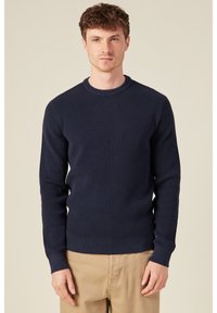 Maglione lavorato a maglia blu navy con scollatura rotonda e polsini a coste. Tessuto texture con una vestibilità aderente, indossato con pantaloni beige.