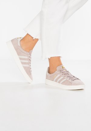 Person iført lyserøde Adidas-sneakers i ruskind med hvide striber og hvide bukser med frynset kant, hvor den ene fod er let løftet.