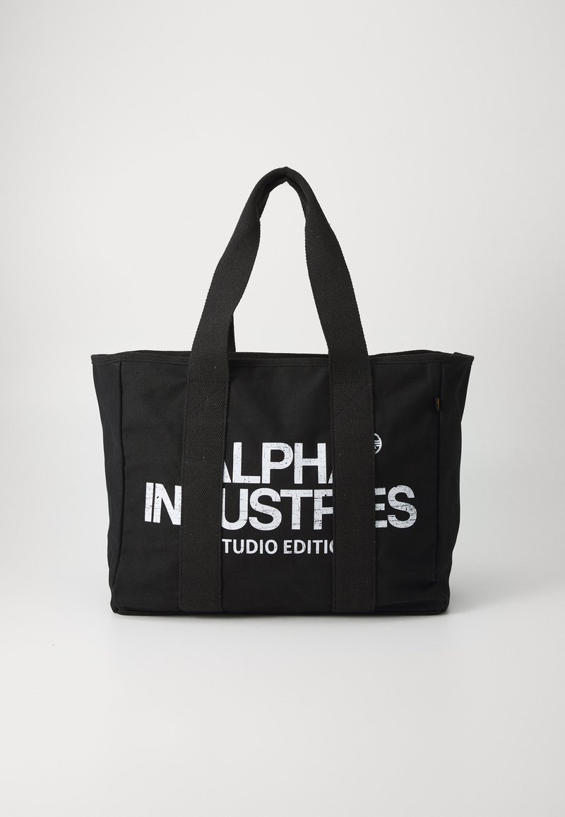 Bolsa de lona negra con dos asas resistentes, que presenta un destacado texto blanco "ALPHA INDUSTRIES STUDIO EDITION" en un lado.