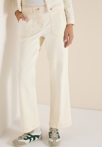 Personne portant un jean crème à jambes larges, des chaussures blanches décontractées avec des accents verts et des chaussettes claires, debout contre un mur et un sol beige.