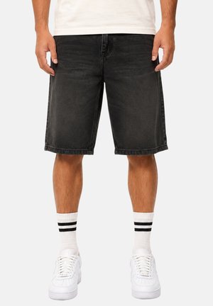 Mann trägt schwarze Denim-Shorts, weiße Sportsocken mit zwei schwarzen Streifen und weiße Turnschuhe, steht vor einem einfarbigen Hintergrund.