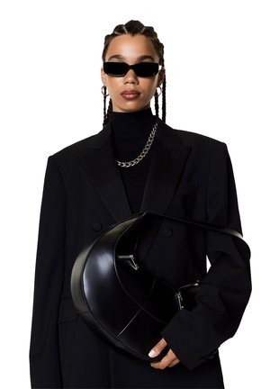 Blazer noir ajusté sur un col roulé noir, accompagné de bijoux en chaîne argentée. Tient un sac à main noir brillant et courbé avec des zips.