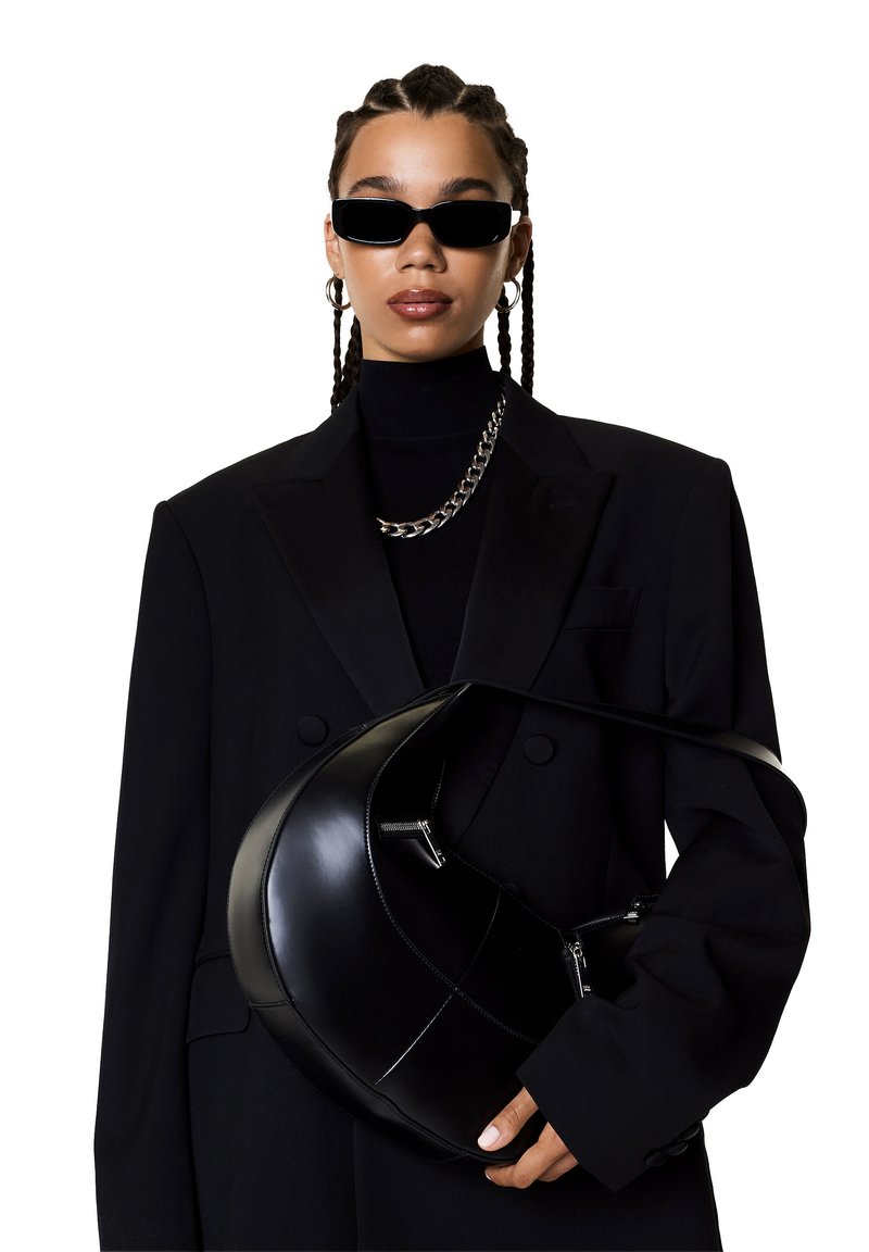 Blazer noir ajusté sur un col roulé noir, accompagné de bijoux en chaîne argentée. Tient un sac à main noir brillant et courbé avec des zips.