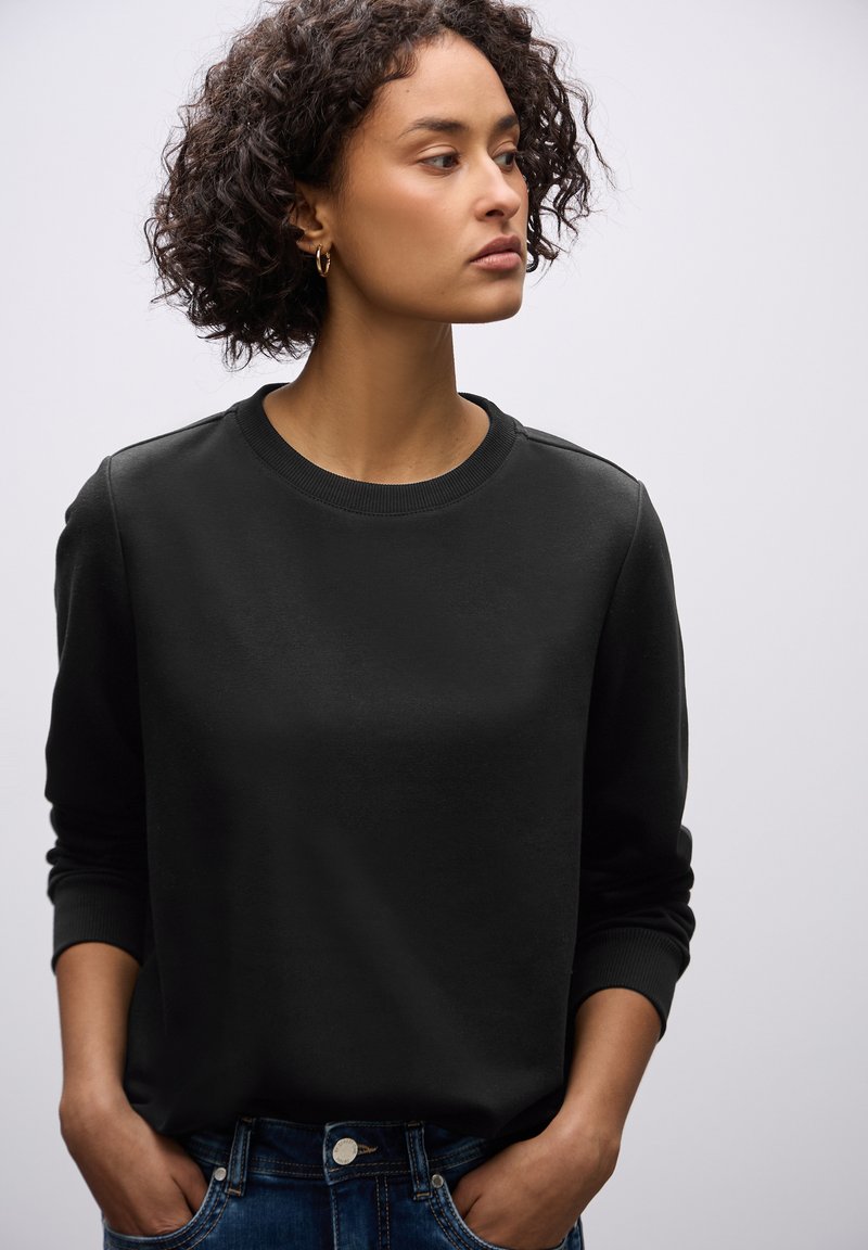 Sudadera negra con cuello redondo, puños acanalados y una textura suave, combinada con jeans de mezclilla azul.
