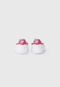 Balta Nike sporta apavu pāris ar sarkaniem papēža akcentiem un logo, fotografēts no aizmugures uz vienkārša balta fona.