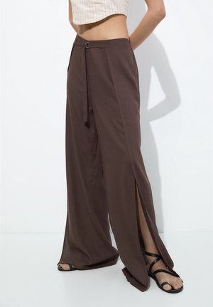 Donna con pantaloni marroni a gamba larga con spacchi laterali, cintura annodata in vita e sandali neri con cinturini, in piedi davanti a uno sfondo bianco.