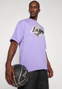 Tricou scurt de culoare lavandă cu mânecă scurtă, prezentând logo-ul "Los Angeles Lakers" și o grafică cu o minge de baschet; purtat cu pantaloni scurți din plasă negri și ținând o minge de baschet Spalding.