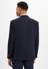 Granatowa marynarka z teksturowanym wykończeniem, z pojedynczym rozcięciem z tyłu i srebrnymi guzikami na rękawach, noszona na ciemnych spodniach.