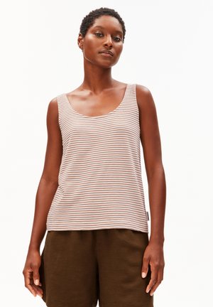 Frau mit kurzen Haaren, die ein beige-weiß gestreiftes ärmelloses Top und dunkelbraune Shorts trägt, steht vor einem weißen Hintergrund.