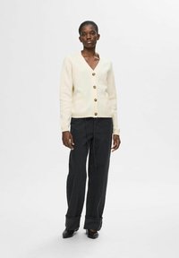 Cárdigan de punto color crema con escote en V, cinco botones y dos bolsillos frontales, combinado con jeans de pierna ancha negros y zapatos negros.