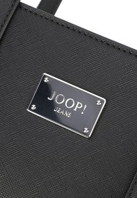Črna teksturirana platnena torba z metalizirano srebrno oznako JOOP! JEANS, okrašena s štirimi okrasnimi zakovicami. Enostaven dizajn brez vidne kovinske opreme.