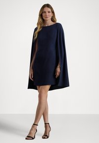 Tumši zila mini kleita ar apmetņa piedurknēm, izgatavota no gludas auduma. Ir laivas formas kakla izgriezums un saskaņota ar melnām kurpēm ar siksniņām.