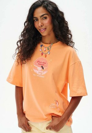 Kobieta o długich kręconych włosach, ubrana w pomarańczowy T-shirt z grafiką syreny i warstwowymi, kolorowymi naszyjnikami z koralików na białym tle.