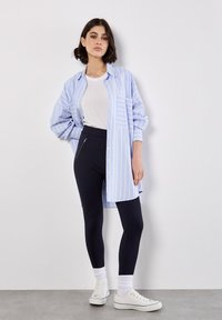 Jasnoniebieska i biała w paski oversize'owa koszula, biała koszulka, czarne legginsy i białe sneakersy z prążkowanymi skarpetkami. Koszula zapinana ma kieszeń z przodu.