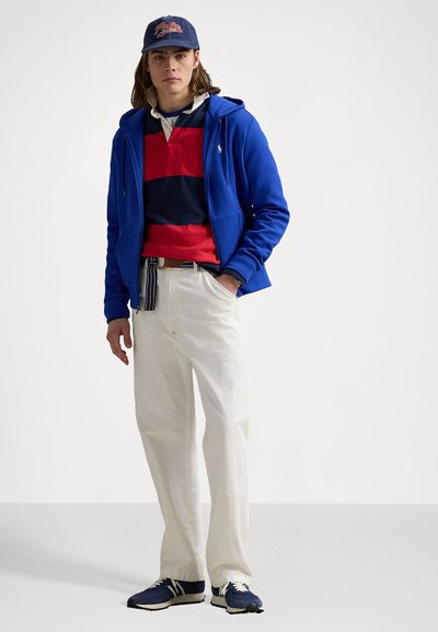 Jovem com um boné azul, casaco azul royal com fecho éclair, camisa de rugby listrada em vermelho e azul-marinho, calças brancas e ténis azul-marinho, de pé com as mãos nos bolsos.