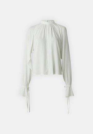 Blusa blanca con cuello alto, detalles fruncidos y mangas largas. Las mangas tienen puños acampanados con lazos. Tejido suave y ligero.