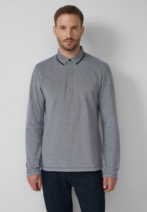 Homme portant un polo à manches longues gris avec un col à bord foncé et un pantalon foncé, se tenant contre un fond clair et uni.