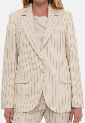 Donna che indossa giacca da completo crema a righe sottili e pantaloni coordinati con dettagli sottili in paillettes, abbinati a una maglia beige sotto.