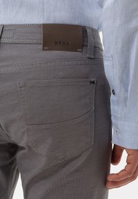 Pantaloni in tessuto grigio con una patch in pelle marrone BRAX sul retro. Caratteristiche includono due tasche posteriori e un motivo testurizzato sottile.