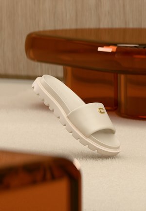 White slide sandal with a thick ridged sole and gold emblem, positioned on light carpet near translucent brown furniture.