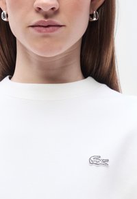 Sweatshirt blanc avec un petit logo crocodile brodé sur le côté gauche de la poitrine. Texture lisse avec un col rond classique.