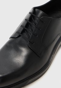 Zapato negro de cuero con cordones, de textura suave, puntera redondeada y suela resistente. Presenta seis ojales y acentos cosidos para mayor durabilidad.