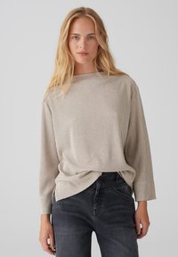 Helles beiges, geripptes Langarmoberteil mit Bootsausschnitt und lockerer Passform, kombiniert mit dunkelgrauen Jeans. Die Textur ist weich und leicht strukturiert.