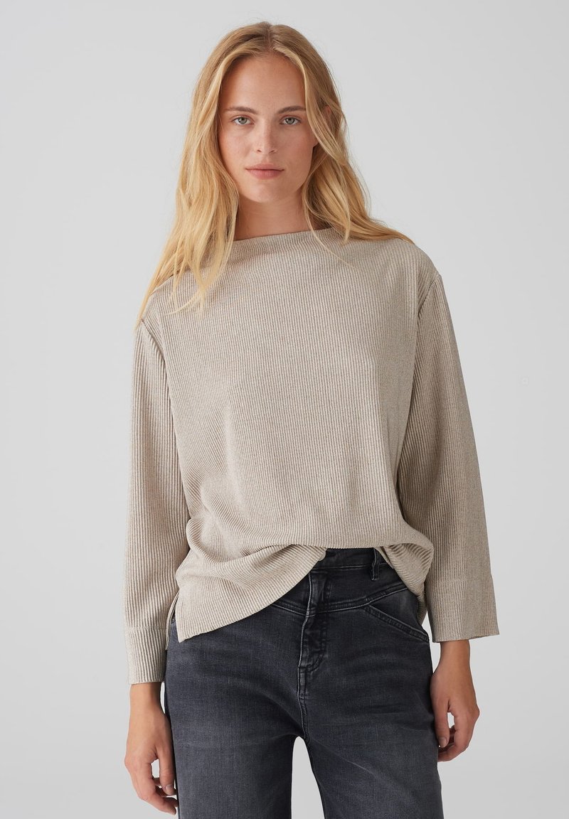 Helles beiges, geripptes Langarmoberteil mit Bootsausschnitt und lockerer Passform, kombiniert mit dunkelgrauen Jeans. Die Textur ist weich und leicht strukturiert.