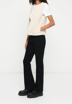 Frau trägt eine beige ärmellose, flauschige Weste über einem weißen kurzärmeligen Hemd, schwarze Hosen und schwarze klobige Plateauschuhe, steht seitlich.