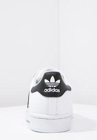 Valkoiset tennarit, joissa on nahkainen päällinen, musta kantapaneli, jossa on Adidas-logo, litteä valkoinen pohja, eikä näkyviä kuvioita tai korostuksia.