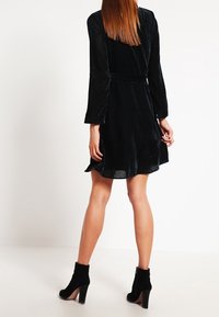 Robe en velours noir avec des côtes verticales, manches longues et taille cintrée ; associée à des bottines à talons noires.