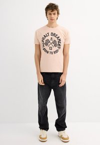 Camiseta de manga corta rosa con un diseño gráfico negro que incluye banderas a cuadros y texto. Combinada con jeans negros y zapatillas beige.