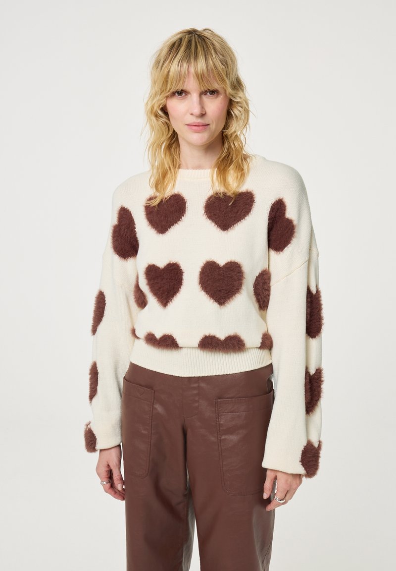 Witte sweater met donzige bruine hartpatronen, losse fit, geribde halslijn en zoom. Draag het met een bruine leren broek.
