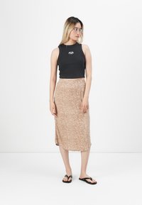 Mujer joven que lleva gafas transparentes, top corto sin mangas negro, falda midi beige con estampado y chanclas negras, de pie contra un fondo blanco.